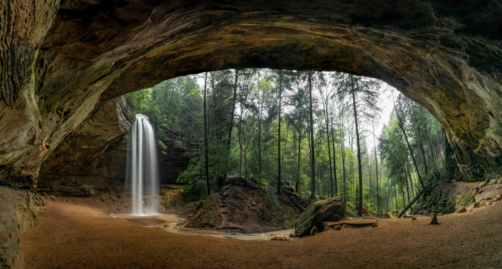 The Ash Cave in Hocking Hills with a waterfall coming over the sides, surrounded by trees. Its one of the best things to do in Ohio with kids. 