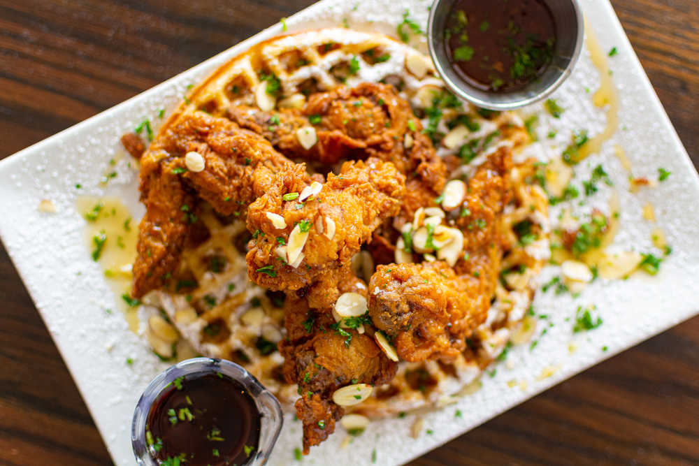 A white plate piled high with waffles and fried chicken that is drizzled in honey, powdered sugar, scallions, and nuts. 
