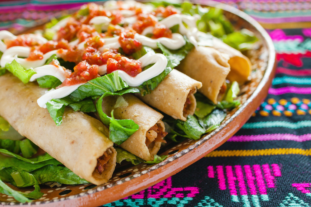 A plate of chicken flautas on a bed of lettuce with salsa and cream cheese on them. The plate is on a colorful patterned table cloth. It's similar to dishes you'd find at one of the best restaurants in Grand Rapids. 