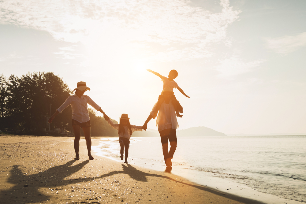 A family with two adults and two kids walking on a sandy beach as the sun is setting in the back behind some trees. 