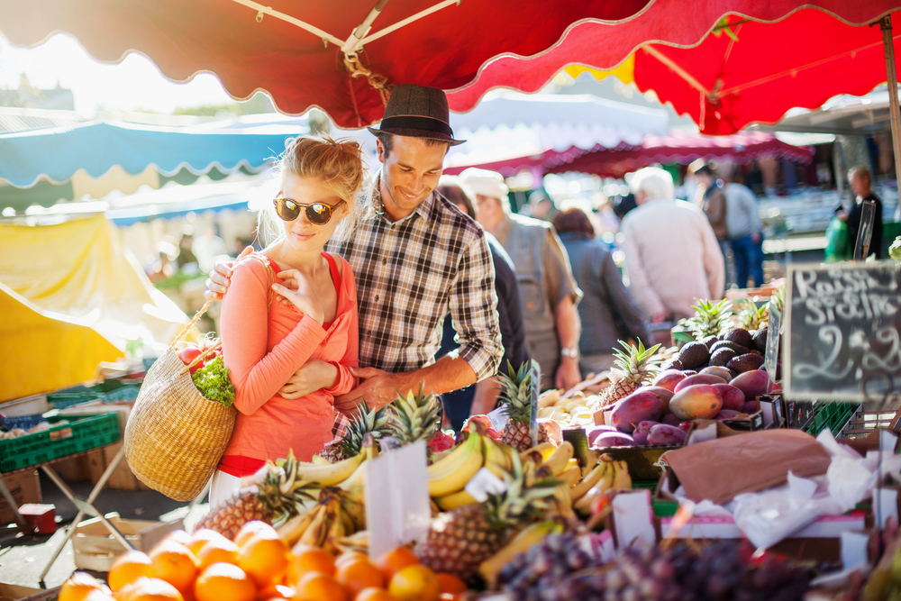 A couple looking at a selection of fruits and vegetables at an outdoor farmers market on a sunny day. 