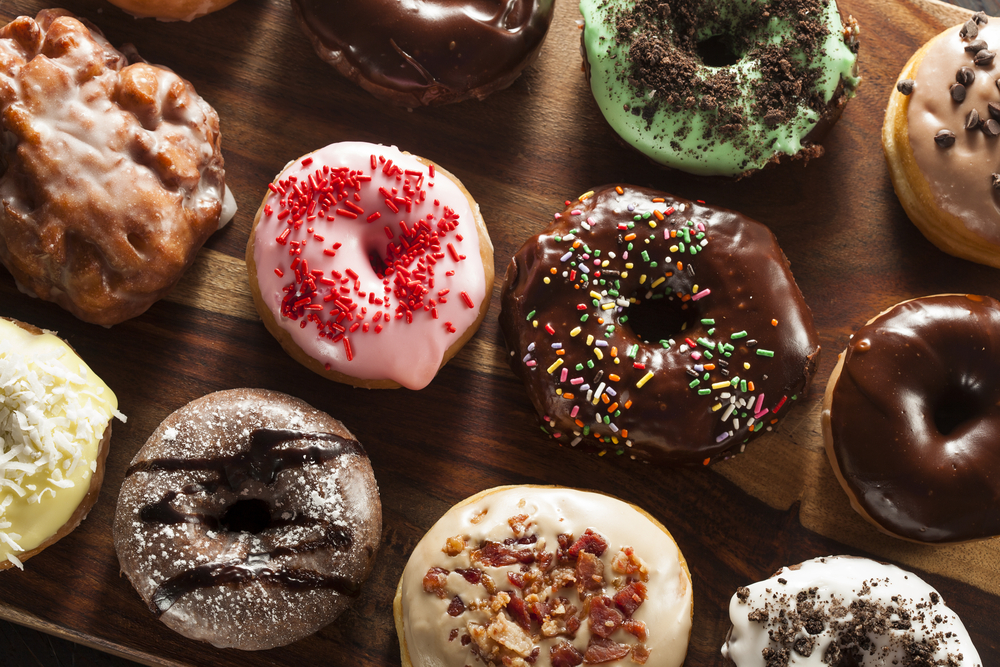A wooden tray full of gourmet donuts with frosting, sprinkles, and other toppings. 
