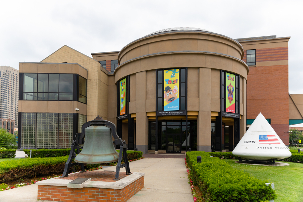 The exterior of the Grand Rapids Public Museum. It is a brick building with a bell and a replica of the Apollo space shuttle in front of it. 