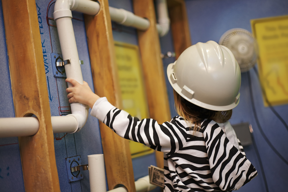 A kid wearing a white hard hat interacting with a plumbing exhibit in a children's museum. 