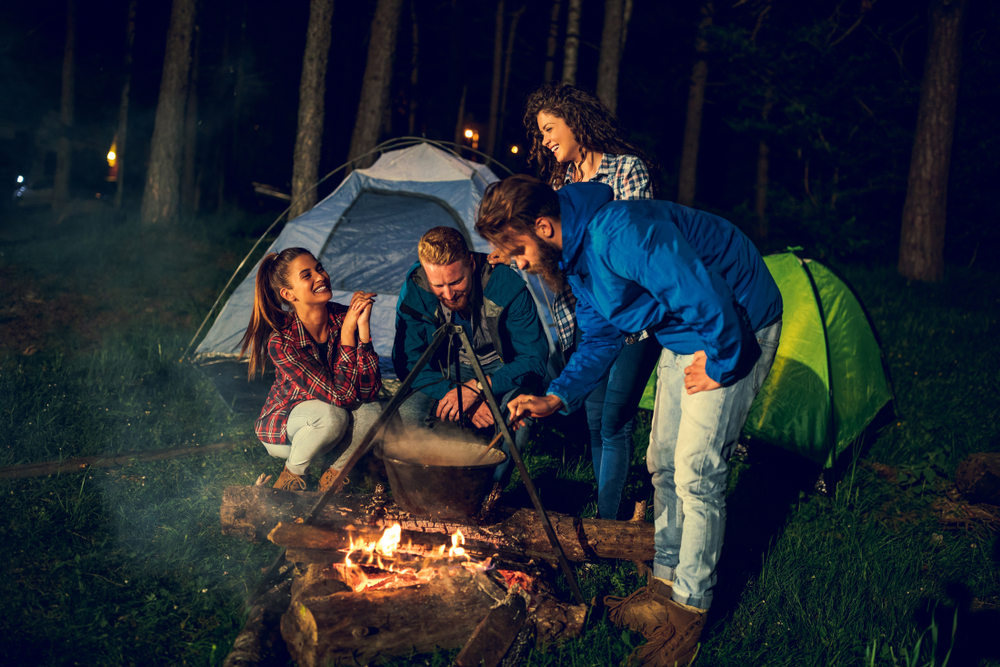 A group of people standing around a campfire at night with a big kettle hanging over it, it's similar to camping in the UP.