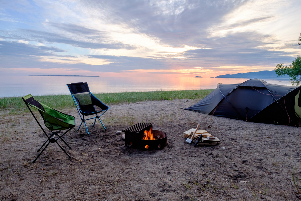 A tent and two chairs near a bone fire in the sand next to a lake.
