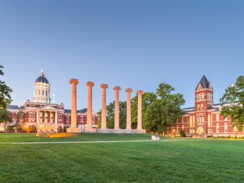 A view of two brick buildings with towers and a series of columns on a lawn. It is part of the University of Missouri campus, one of the best things to do in Columbia MO