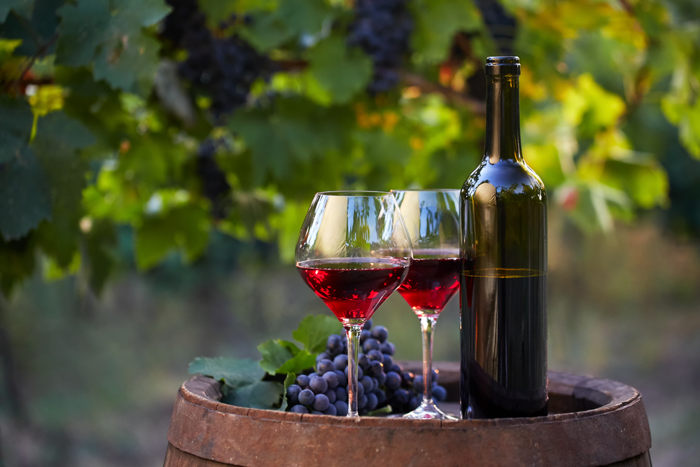 Two glasses of red wine on a wooden barrel with bottle to right and greenery in background wineries in Indiana