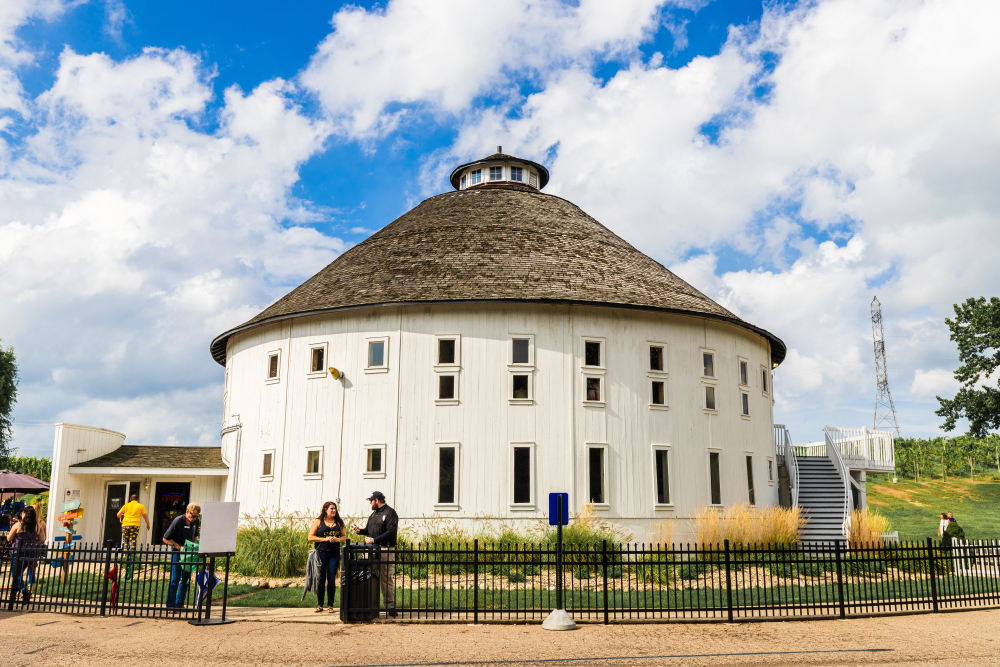 Round Barn Winery, Distillery & Brewery features a white barn that houses their tasting room and shop. The barn is a large white barn and there is greenery and a fence around it.  