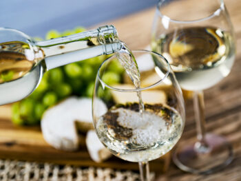 Glass of white wine being poured into wineglass with cheese, grapes and second glass of white wine in background at one of the wineries in Wisconsin