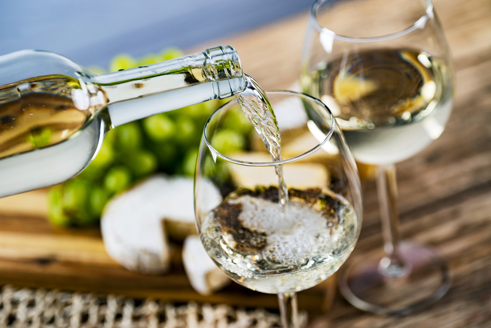 White wine been poured into a glass with grapes and cheese in the background. 