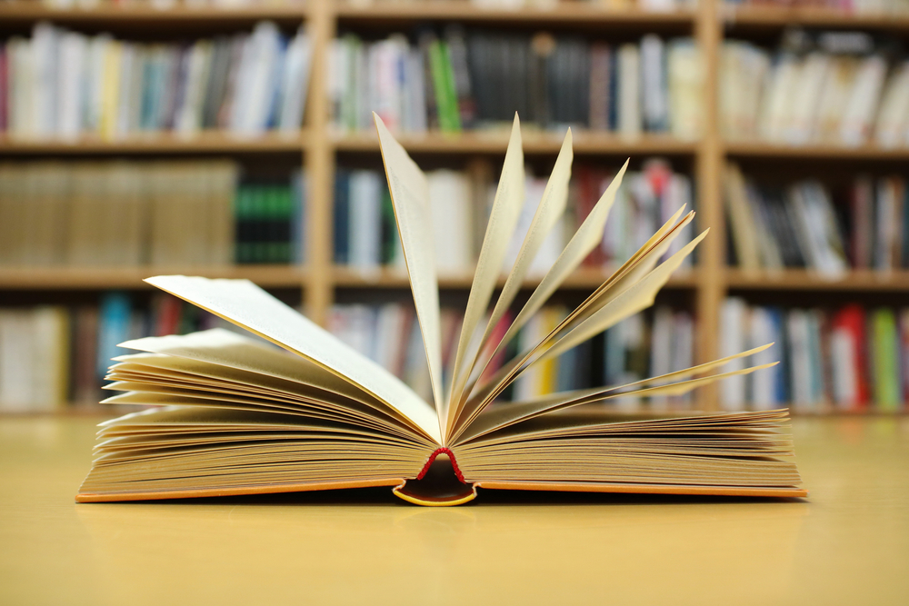 An open book on a table with a bookcase in the background.