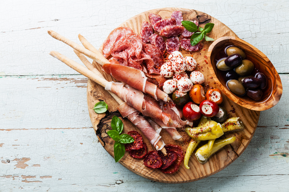 A tray of antipasto on a white wooden table similar to what you'll find at restaurants in Dayton. The tray has olives, cheese, cured meats, peppers, and bread sticks. 