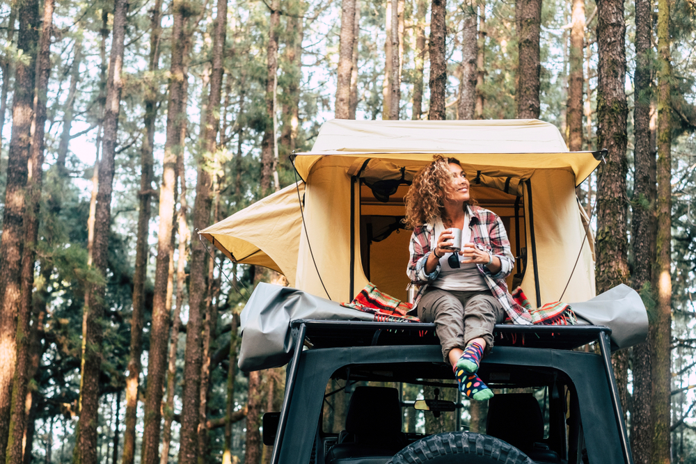 Car with tent on the roof and young woman sitting down outside looking at the forest around. She has a cup in her hand. 
