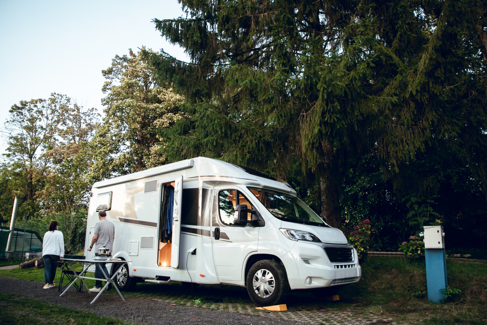 White motor home parked in camping in the woods next to large trees and two people coming out the door of the home