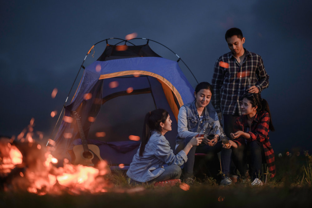 Group of friends sitting outside a tent in the dark. There is a fire in the foreground. 