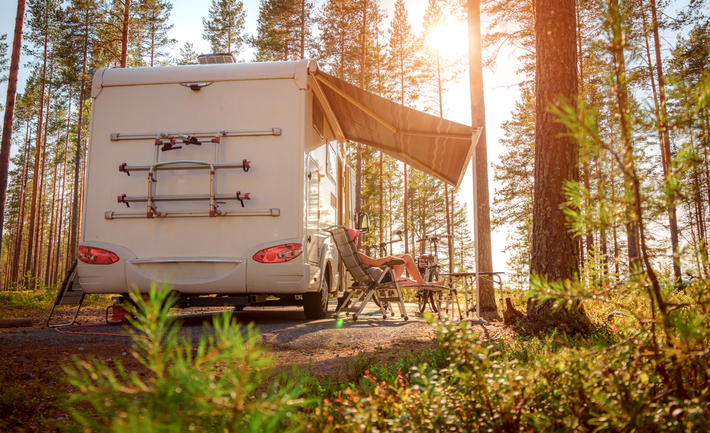 A motorhome with an awning up and chairs outside. The RV is in the woods. 