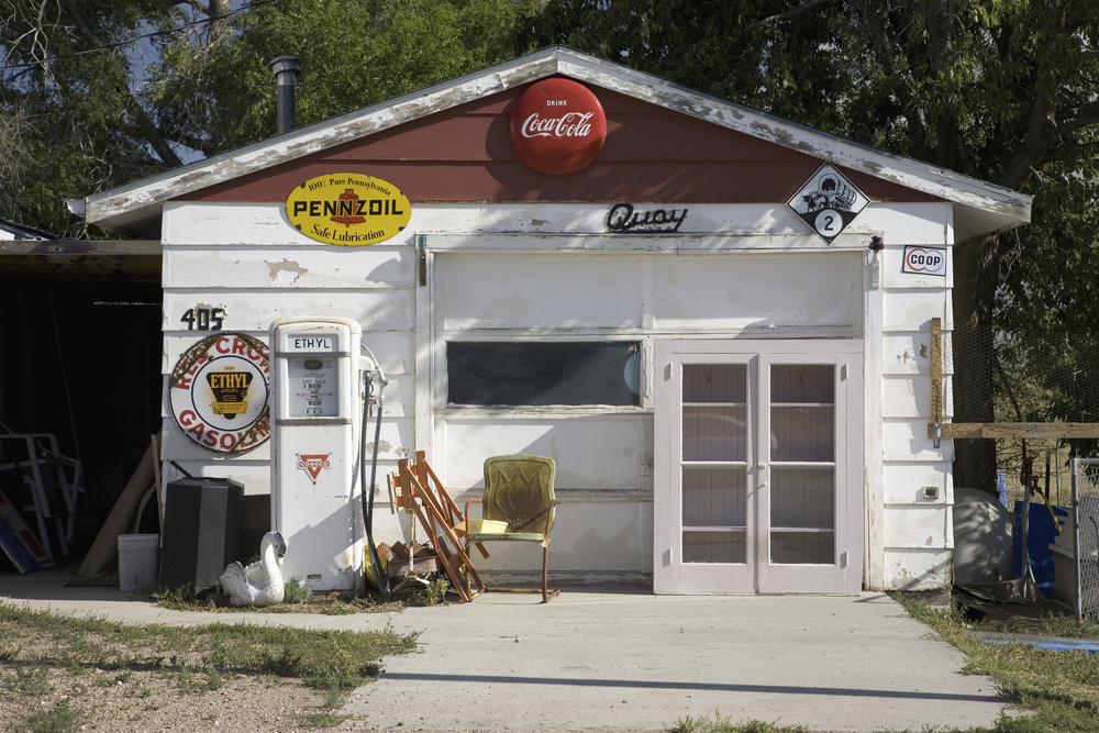 A small rural gas station that is white with only one pump that is no longer in use. The building has old gas signs and drink signs on it. 