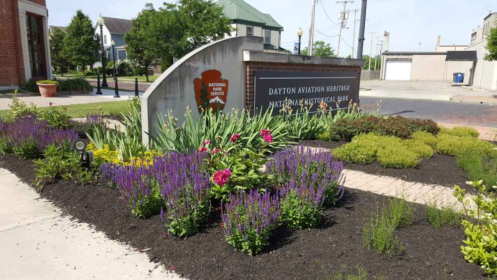 The sign at the entrance of the Dayton Aviation Heritage National Historic Park which has purple, pink, and yellow flowers and shrubs around it. 