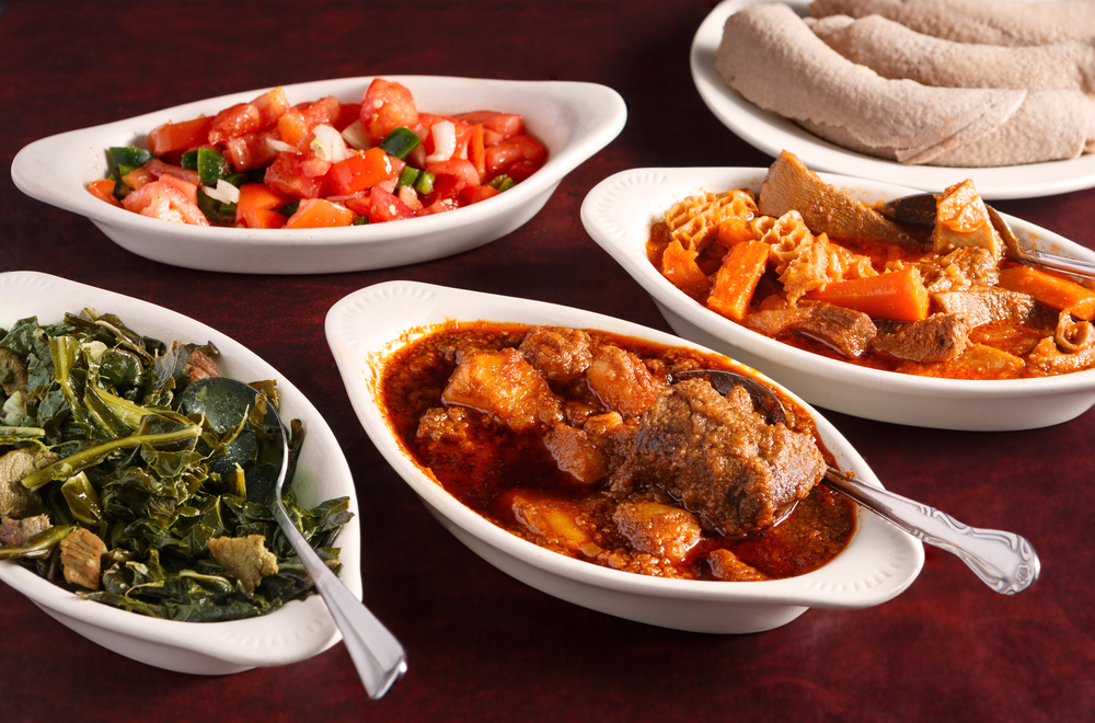 A variety of white trays full of different Ethiopian foods like stewed meats, tomato salad, and cooked greens. Its similar to food you'll find at restaurants in Dayton . 