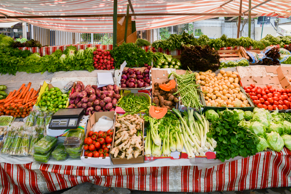 A stand with fresh produce at an open-air market with red and white fabric hanging over it and on the stand. 