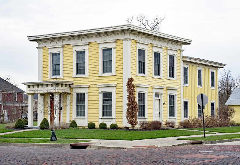 A historic Italianate style yellow home on a street with a brick road. 