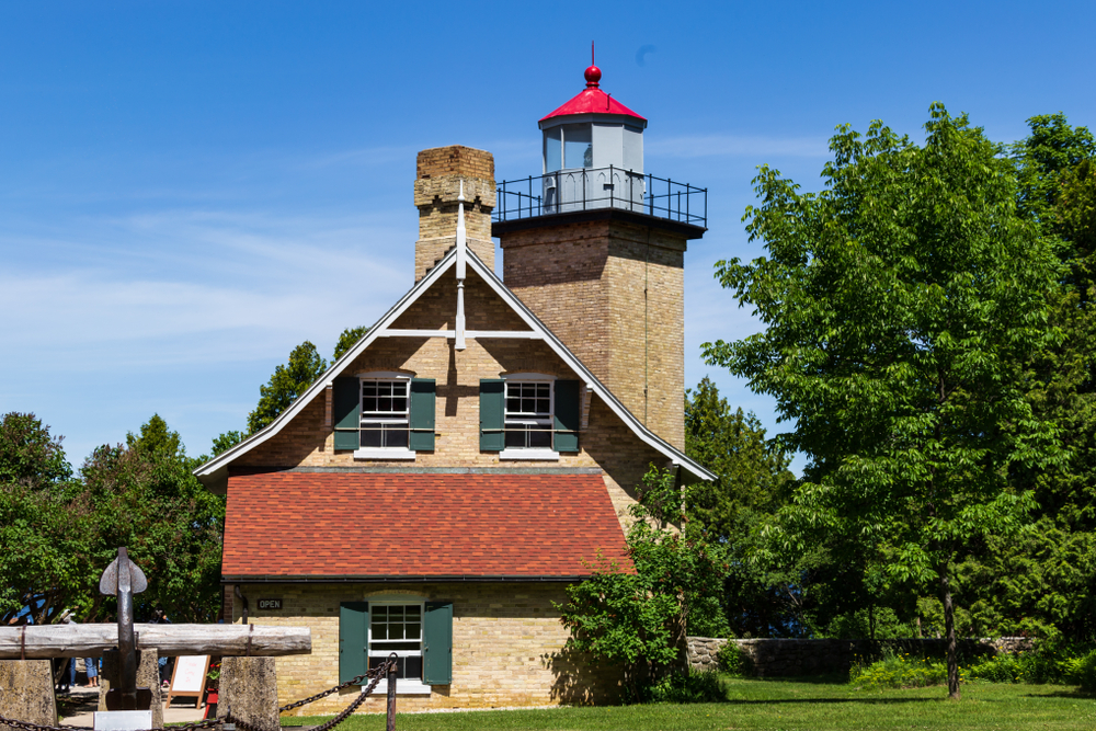 Eagle Bluff Lighthouse is a brown building with a tower behind it. It is surrounded by trees. 