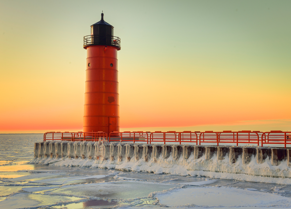 A red lighthouse with a pier with a red fence around. Its at sunset and the sky is red and orange. The article is about lighthouses in Wisconsin. 