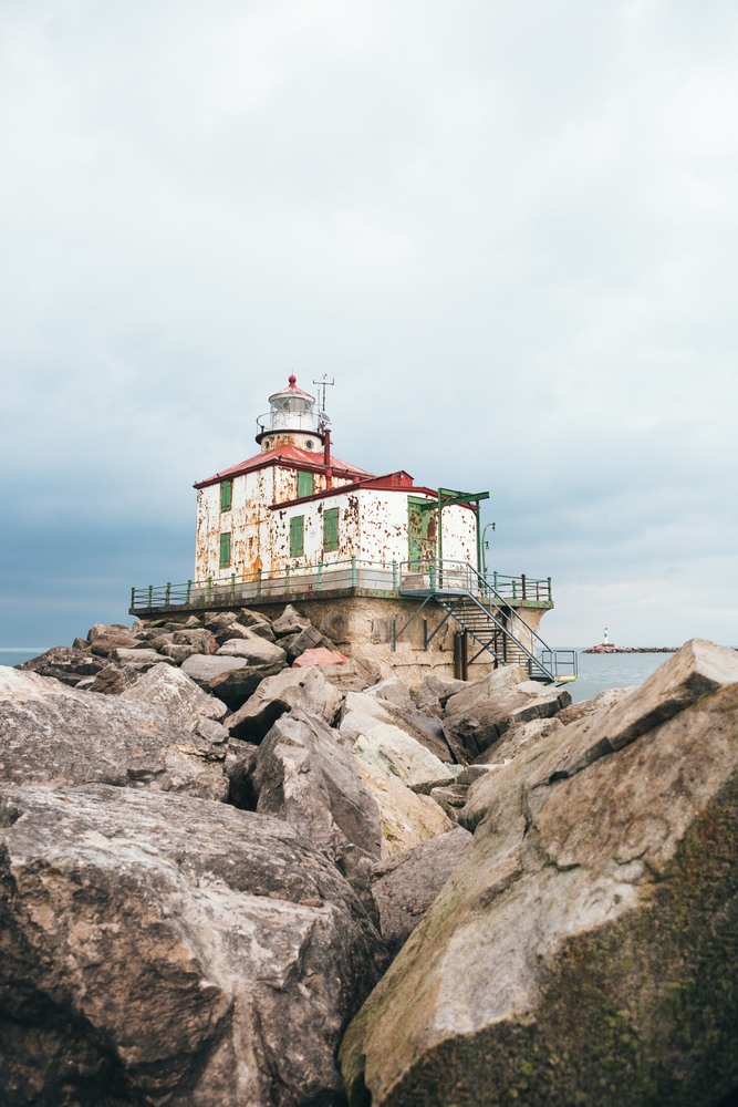 A run down white box style lighthouse. It has green window and door coverings and stands atop a platform and cliffs. Its one of the lighthouses in Ohio.   