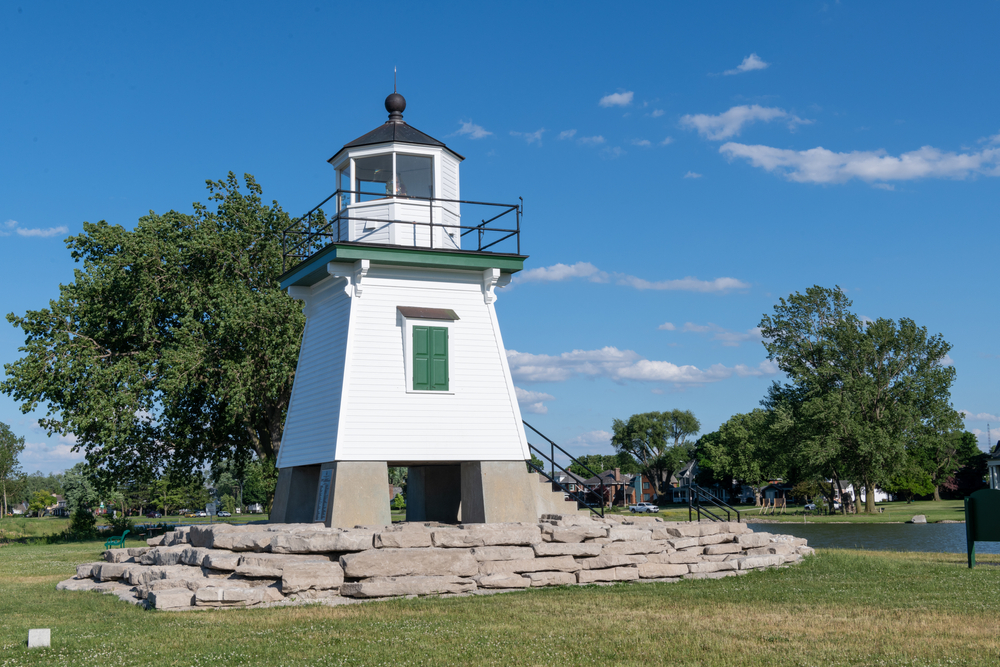 A white lighthouse stands on a grey plinth. It is in a park surrounded by trees.  