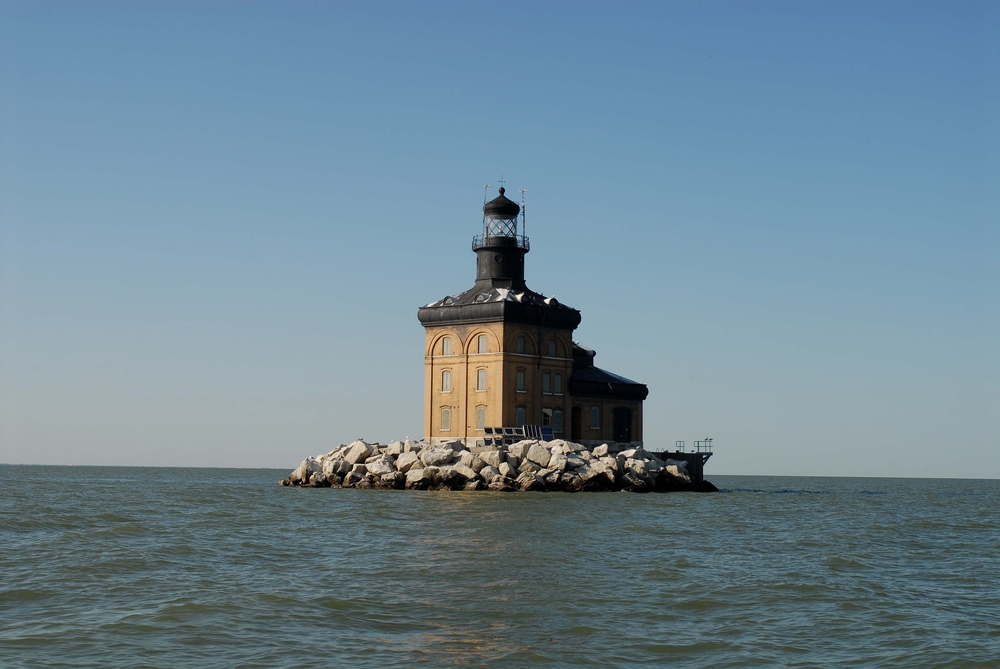 The brown lighthouse has a black roof and it made of two buildings. Its on a stone outcrop with a black light on top. 