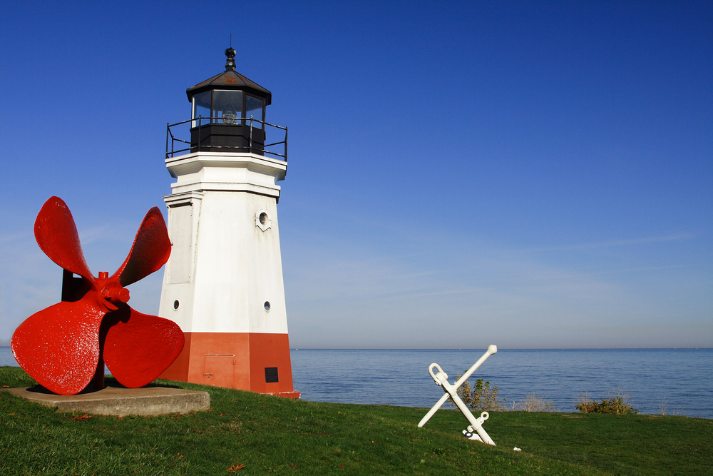 A lighthouse on the shore with the sea behind. The lighthouse in white with red at the bottom. It has a large propellor next to it and an anchor in front. 