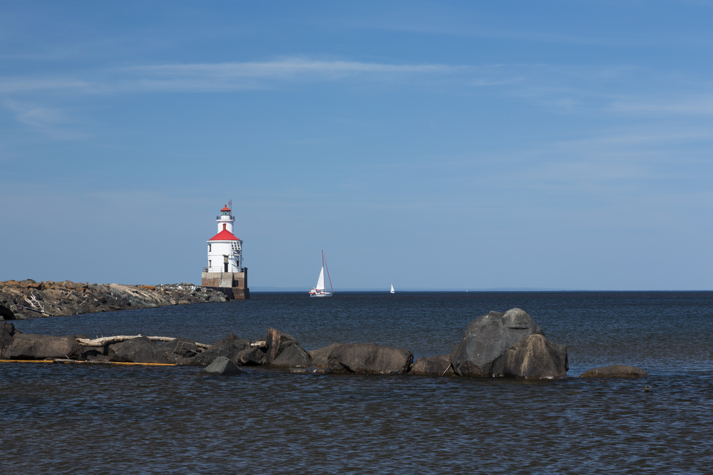 Wisconsin Point Lighthouse out on a rocky outcrop. It's a white lighthouse with a red roof. There is a sailing boat on the water . It's one of the best lighthouses in Wisconsin.