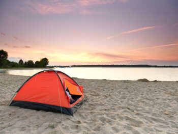 An orange tent on the sandy shores of a lake which is a great place to go camping in Illinois. The sun is starting to set over the lake.