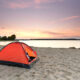 An orange tent on the sandy shores of a lake which is a great place to go camping in Illinois. The sun is starting to set over the lake.
