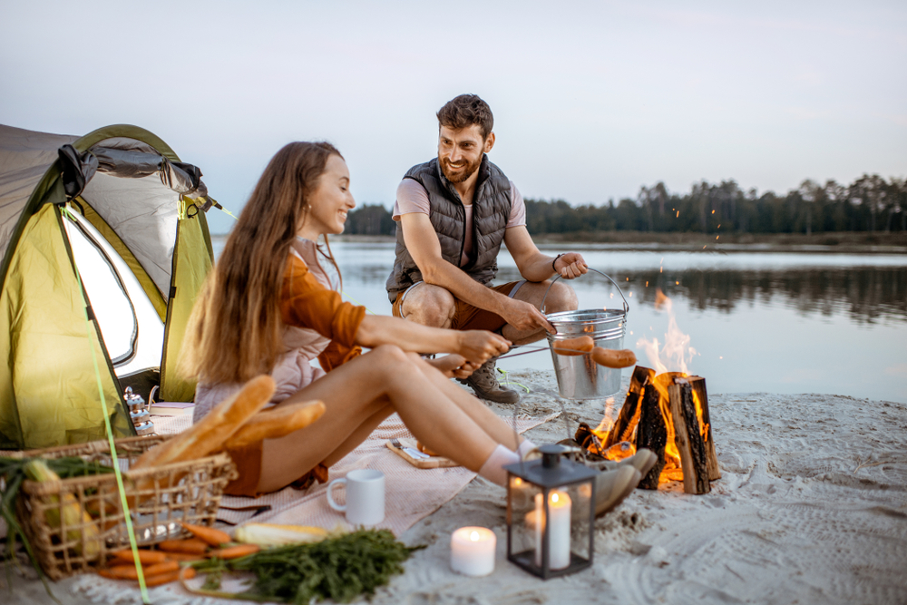 A couple camping on a sandy beach with a tent. They are roasting food over a small bonfire. 