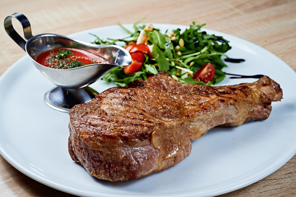 Grilled rib steak with greens salad and tomato sauce on a white plate