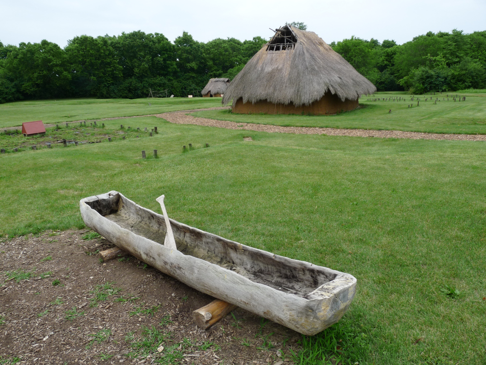 A hand carved canoe, a building with a thatched roof, and a grassy meadow at a archeological park in Dayton Ohio. 