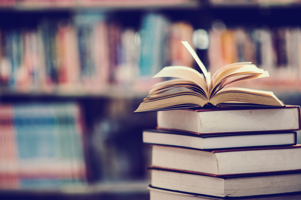 Books piled up on a table in a library. One of the books is open and the pages are fanned out. 