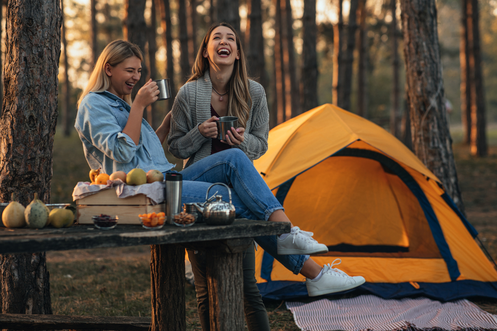 Two women laughing as they enjoy a hot beverage outside of a yellow tent in the middle of the woods. 