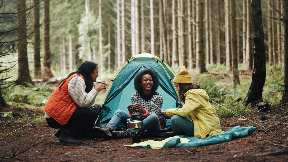 Three women sitting around a kettle in front of a small tent in the middle of the woods. 