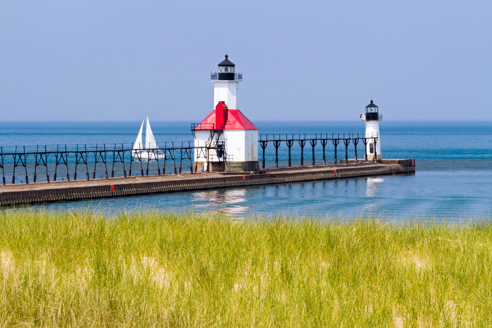 View across a grassy dune to St. Joseph's North Pier and two lighthouses with a sailboat in the lake nearby.