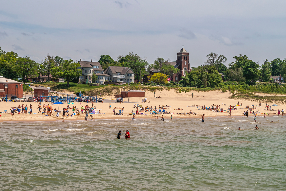 Dark brown stone Saint Basil Catholic church behind beach on Lake Michigan. Playing people and belt of green foliage.