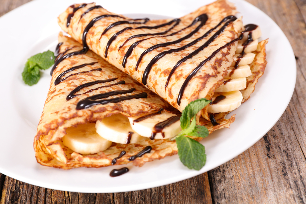 A white plate with crepes that have bananas in them and chocolate drizzled on top. Its similar to what you can get at one of the many restaurants in Bloomington.