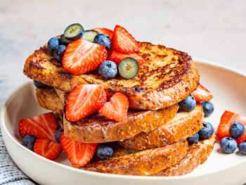Pile of French toast with strawberries and strawberries on top of it and in the white bowl.