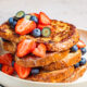 Pile of French toast with strawberries and strawberries on top of it and in the white bowl.