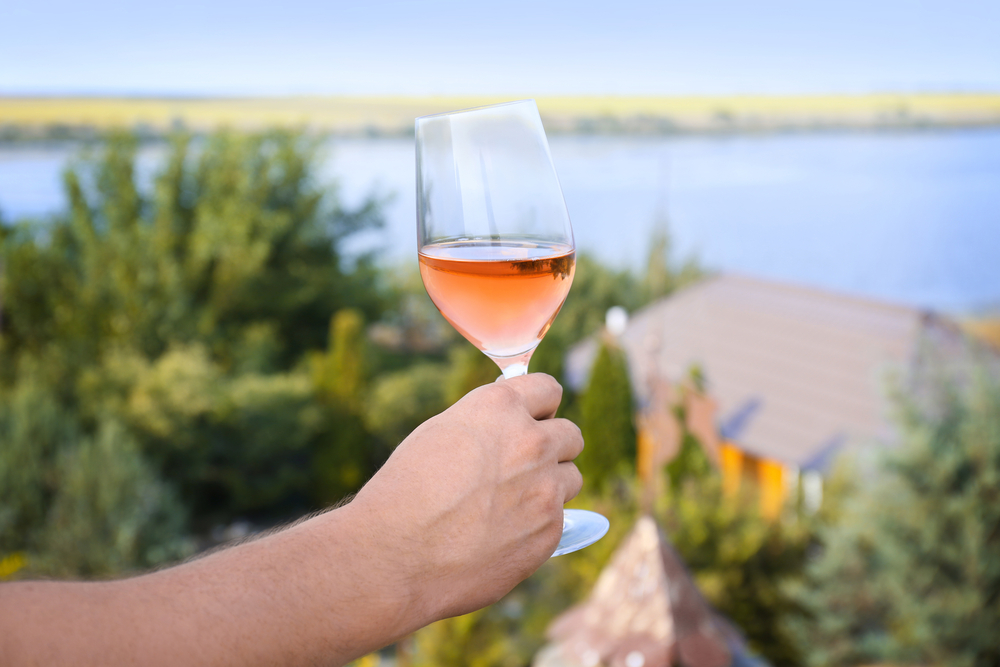 A Caucasian person holding a glass of rose with trees, a building, and a river in the background wineries in Missouri