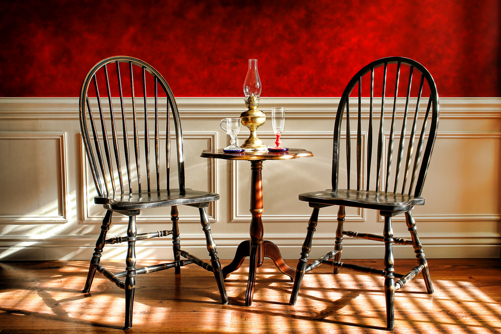 A set of chairs and a small table on display in a historic house museum. The wall behind the chairs has a chair rail and the top is a textured red wallpaper. 