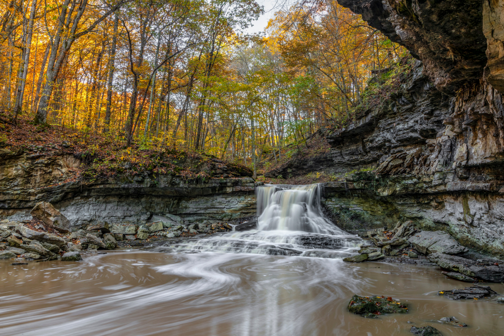 A small waterfall surrounded by limestone rocks and a dense forest in the fall. The leaves on the trees are yellow, orange, and green. 