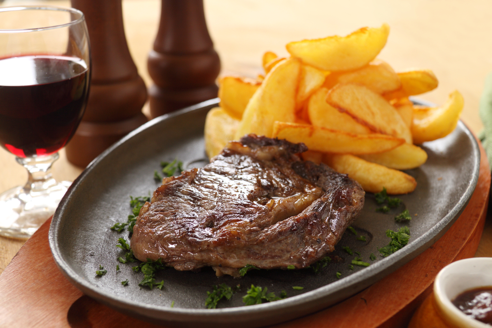 Grilled rib fillet steak with fried chips on a plate with a glass of red wine on the side. The article is about restaurants in Bay City. 